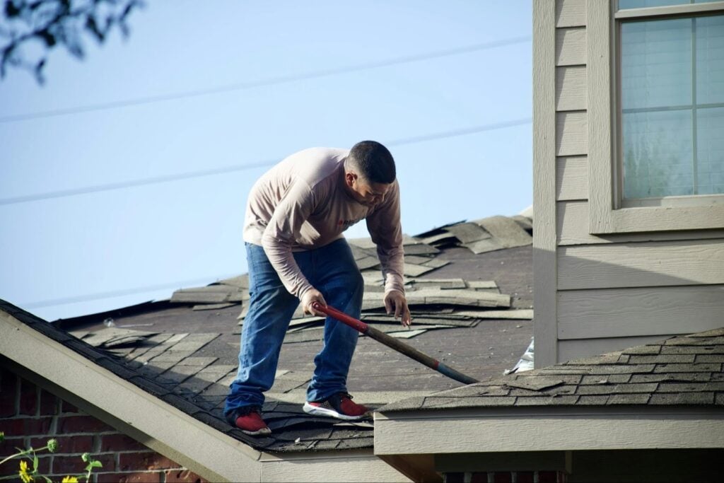 Roofer removing damaged shingles during an urgent roof repair, showing the type of situation where homeowners should request emergency roof leak repair services.
